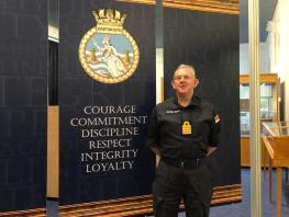 Principal Paul Little, Honorary Captain Royal Navy Reserves wearing his uniform standing in front of a wall with an insignia and 